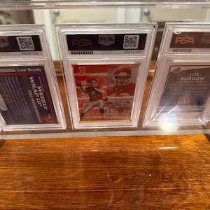 May include: A clear acrylic display stand holding three graded sports cards. The cards feature images of football players Tom Brady and Joe Burrow. The stand has silver metal supports and is set on a wooden surface.