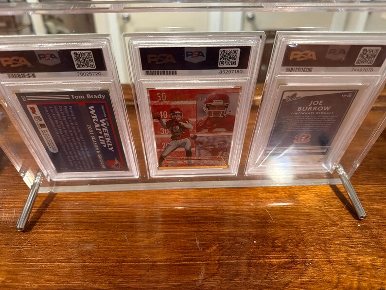 May include: A clear acrylic display stand holding three graded sports cards. The cards feature images of football players Tom Brady and Joe Burrow. The stand has silver metal supports and is set on a wooden surface.