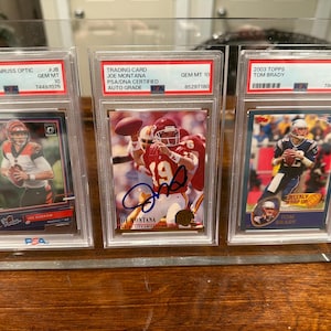 May include: Three encased football trading cards featuring Joe Burrow, Joe Montana, and Tom Brady. The cards are in clear protective cases, with the Joe Montana card featuring an autograph. The cards are from the 2020 Panini Donruss Optic, 2003 Topps, and a Joe Montana trading card.