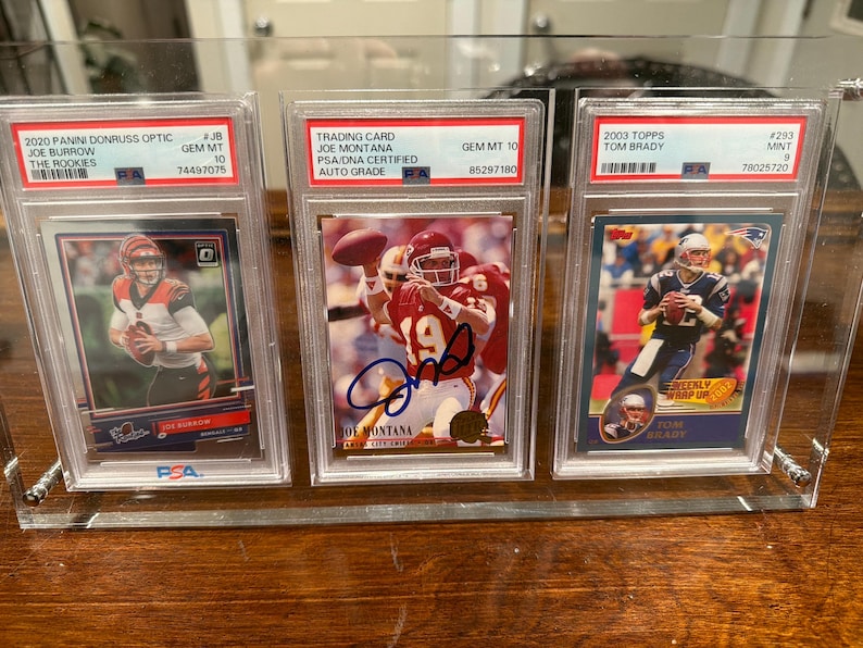 May include: Three encased football trading cards featuring Joe Burrow, Joe Montana, and Tom Brady. The cards are in clear protective cases, with the Joe Montana card featuring an autograph. The cards are from the 2020 Panini Donruss Optic, 2003 Topps, and a Joe Montana trading card.