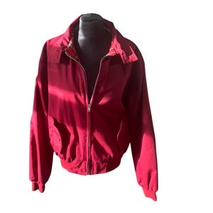 Può includere: Giacca rossa con chiusura a zip completa. La giacca ha un collo e due tasche anteriori. La fodera interna presenta un motivo a quadri. La giacca è esposta su una testa di manichino nera.