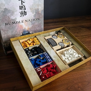May include: Open board game box with the title "Rumble Nation" visible. The box contains various game pieces in yellow, black, blue, and red, including dice and tokens. The game appears to be strategy-based.