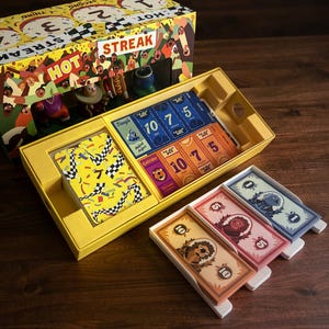 May include: A board game box set with a yellow exterior and the word "Streak" in red. The box is open, revealing game cards and components. The cards feature colorful designs and numbers. The game's theme appears to be racing.