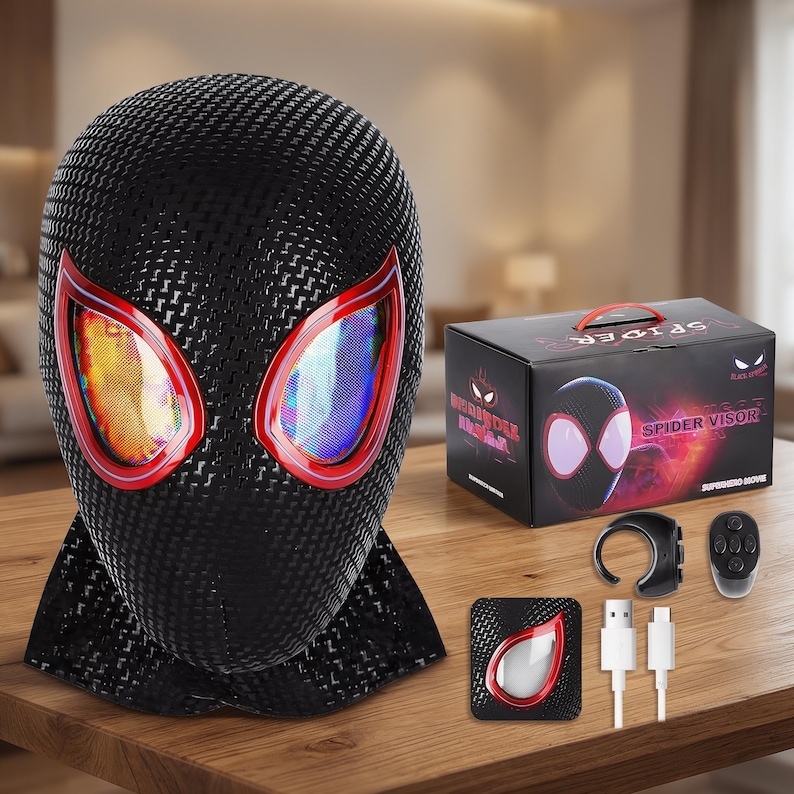 May include: A black Spider-Man visor with red-rimmed eye lenses. The visor is made of a textured material and sits on a wooden surface. A box and accessories are also present, including a remote and charging cables.