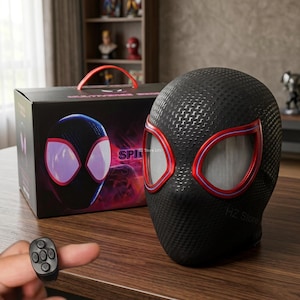 May include: Black speaker shaped like a superhero mask with red and white accents. The speaker has a textured surface and is next to a box with a matching design. A small remote control is in the foreground.