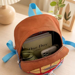 Puede incluir: Una mochila marr&oacute;n con correas azul brillante y un bolsillo frontal amarillo con patr&oacute;n de cuadr&iacute;cula. El compartimento principal abierto contiene un cuaderno con el texto One Day en la portada, un tel&eacute;fono inteligente blanco y un objeto de tela verde oliva.