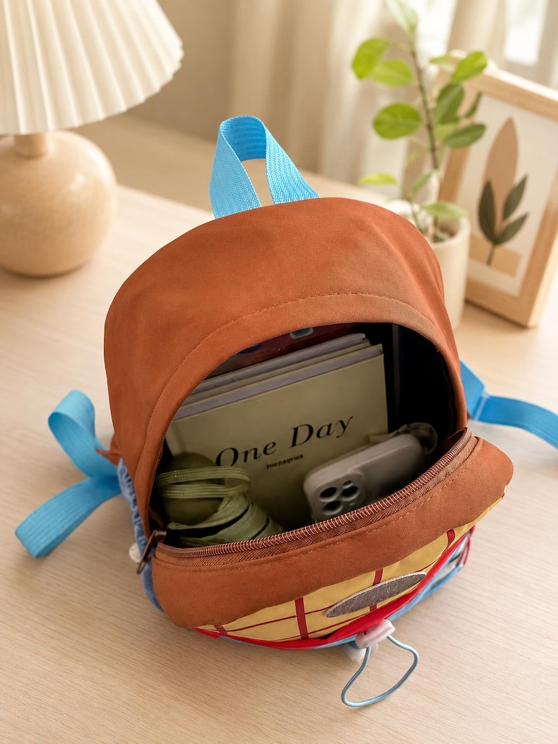 Puede incluir: Una mochila marr&oacute;n con correas azul brillante y un bolsillo frontal amarillo con patr&oacute;n de cuadr&iacute;cula. El compartimento principal abierto contiene un cuaderno con el texto One Day en la portada, un tel&eacute;fono inteligente blanco y un objeto de tela verde oliva.