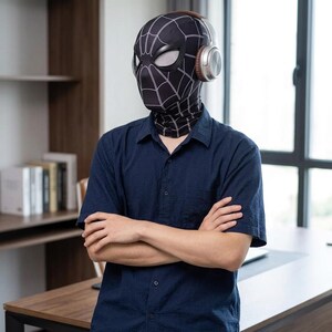 May include: A black Spider-Man mask with white web detailing, worn with silver headphones. The mask covers the head and neck. The person is wearing a dark blue short-sleeved button-down shirt.