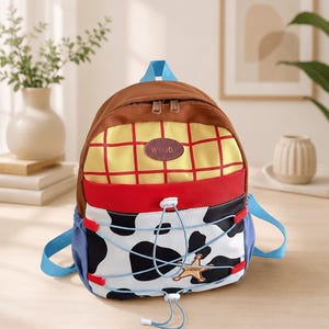 Puede incluir: Una mochila con parte superior marr&oacute;n, una secci&oacute;n amarilla con patr&oacute;n de cuadr&iacute;cula y un parche ovalado marr&oacute;n con la etiqueta WOODY, una banda horizontal roja y una secci&oacute;n inferior con estampado de vaca, cord&oacute;n azul y una estrella de sheriff dorada.
