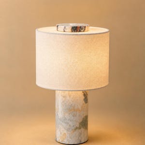 Puede incluir: Una lámpara de mesa moderna con una base cilíndrica de mármol y una pantalla de tela color crema. La lámpara presenta un pequeño acento redondo de mármol en la parte superior. La base tiene tonos grises, verdes y dorados.