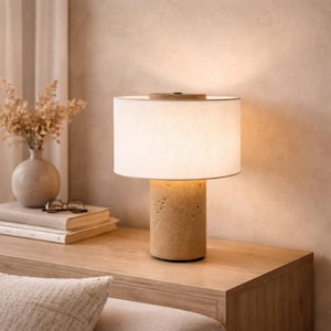 Puede incluir: Lámpara de mesa beige con base cilíndrica y pantalla blanca. La base tiene un aspecto texturizado similar a la piedra. La lámpara está sobre una superficie de madera, con un jarrón de flores secas y libros al fondo.