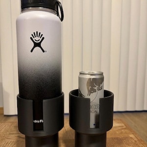 Può includere: Una borraccia Hydro Flask bianca e nera con tappo e logo neri. Accanto, un portabicchieri nero con una lattina argentata. Gli oggetti sono su una superficie di legno.