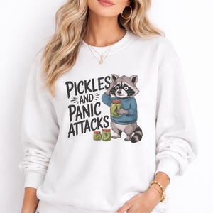 Könnte beinhalten: Weißes Sweatshirt mit dem Text "Pickles and Panic Attacks" und einem Cartoon-Waschbären, der ein Glas Essiggurken hält. Der Waschbär trägt einen blauen Pullover. Zwei weitere Gläser mit Essiggurken befinden sich darunter.