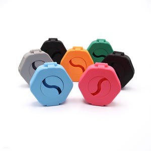 May include: A collection of seven hexagonal cases in various colors including light blue, pink, orange, green, gray, and black. Each case features a hinged lid with a central S-shaped cutout, creating a uniform geometric design across all items.