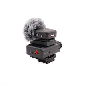 May include: A black wireless microphone receiver unit labeled MIC RX with a fuzzy gray and white windscreen attached to the top. The device features a side-mounted orange port, a link button, and a mounting base for camera attachment.