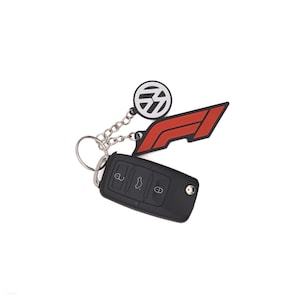 Pode incluir: Um porta-chaves de carro preto preso a um anel de metal prateado. O anel contém dois pingentes: um emblema circular com o logótipo da Volkswagen a preto e branco e um pingente vermelho em forma de logótipo de Fórmula 1 com textura e bordas pretas.