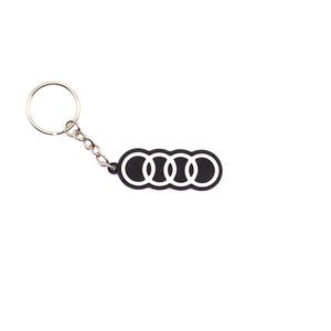 Pode incluir: Um porta-chaves preto e branco com o logótipo dos quatro anéis entrelaçados da Audi. O design consiste numa base retangular preta texturada com quatro contornos circulares brancos, ligada a um anel de metal prateado por uma corrente curta.