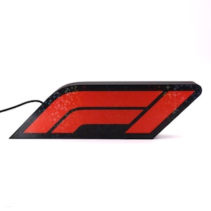 May include: A black and red F1 logo sign featuring a sleek, angular design with a subtle holographic star pattern embedded in the black frame. The bright red central emblem is illuminated, and a thin black power cord extends from the left side.