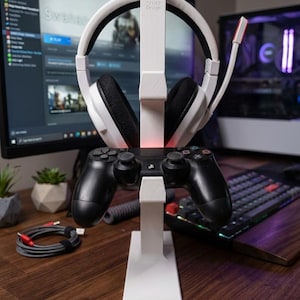May include: A white gaming headset stand with a black controller and a white headset. The stand has the text "TAVAC Design". The headset has black earcups and a microphone. The stand is on a wooden surface.