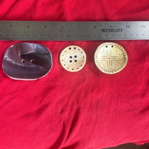 May include: Three buttons positioned below a Westcott ruler. The left button is large, oval, and dark purple with a glossy finish. The middle button is small, round, cream-colored with dots and four holes. The right button is round, cream-colored with a cross design.