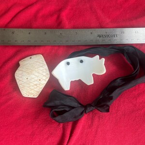 May include: A white, diamond-patterned, shield-shaped pendant and a white, animal-shaped pendant with two black dots, attached to a black ribbon. A Westcott ruler is positioned above the items, showing a measurement of approximately 11 inches.
