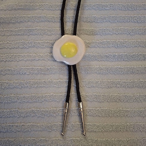 May include: A bolo tie featuring a central ceramic charm shaped like a fried egg with a white oval base and a raised yellow yolk. The charm is attached to a black braided cord that ends in two polished silver-toned metal tips at the bottom.