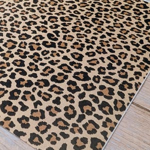 Può includere: Un pezzo di tessuto quadrato con un motivo leopardato. Il motivo presenta macchie nere e marroni su uno sfondo beige. Il tessuto ha un bordo bianco.