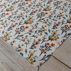 Könnte beinhalten: Ein Stück cremefarbener Stoff mit einem sich wiederholenden Blumenmuster im Aquarellstil. Das Design besteht aus kleinen Blumen- und Blattgruppen in Senfgelb, gedämpftem Orange, zartem Rosa und dunklem Petrol, die in einem Streumuster angeordnet sind.