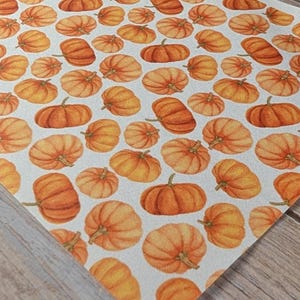 Könnte beinhalten: Ein Bogen Kunstleder mit einem sich wiederholenden Muster aus orangefarbenen Kürbissen auf hellgrauem Hintergrund. Die Kürbisse variieren in Größe und Ausrichtung, jeder mit einem kleinen grünen Stiel und detaillierten Rillen.