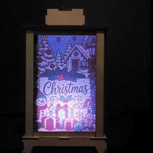 Könnte beinhalten: Ein rechteckiger Holzkasten mit einer hinterleuchteten Illustration einer Winterszene mit Haus, Tannenbäumen, Schneemann, verpackten Geschenken und einer Lebkuchenfigur. Das Wort Christmas ist in einer Schreibschrift in der Mitte zu sehen.