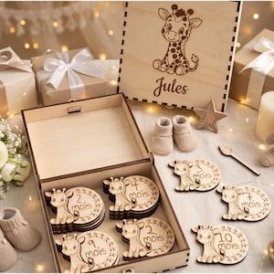 Peut inclure: Une boîte à souvenirs en bois clair avec une girafe gravée au laser et le prénom Jules, contenant des disques de jalonnement en bois avec des illustrations de girafe et des mois en français, accompagnée de chaussons pour bébé beiges.