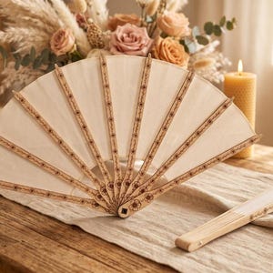 May include: A hand fan with a cream-coloured fabric canopy and light wood ribs. The wooden ribs feature a dark, laser-engraved floral pattern. One fan is fully open in a semi-circular shape, while a second identical fan is folded closed nearby.