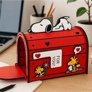 Puede incluir: Un buzón de madera rojo con una figura de Snoopy acostada encima, personajes de Woodstock y corazones. La bandera del buzón es blanca con un corazón rojo y el texto FOR YOU! en negro. El diseño incluye pequeños corazones negros recortados.