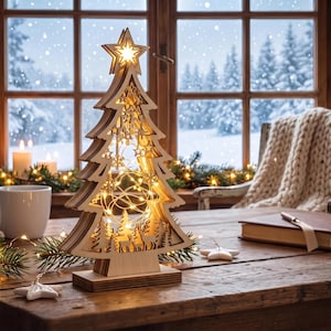 Op de afbeelding: Een gelaagde houten kerstboomdecoratie met een uitgesneden ontwerp van herten en bossilhouetten, verlicht door warme lichtsnoeren en bekroond met een gloeiende ster, staand op een platte rechthoekige houten voet.