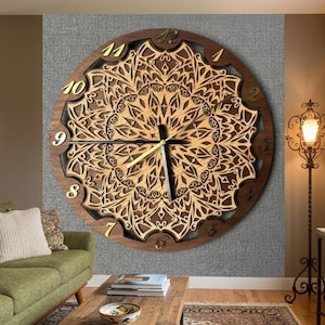 May include: A large circular wall clock featuring a dark brown wooden outer ring and a light tan laser-cut mandala pattern in the center. The clock face displays gold-colored Arabic numerals from one to twelve and thin black clock hands.