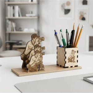 Puede incluir: Un organizador de escritorio de madera clara con un portalápices cuadrado y una base plana. El portalápices muestra una imagen grabada con láser de un fotógrafo. En la base hay tres recortes de madera superpuestos de un fotógrafo con cámara.