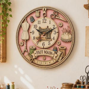 Puede incluir: Un reloj de pared redondo con fondo rosa que presenta ilustraciones de costura, incluyendo un maniquí, tijeras, botones, carretes de hilo, un alfiletero y una cinta métrica. La esfera central muestra una máquina de coser y el texto FAIT MAIN.