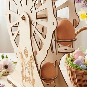 May include: A wooden Ferris wheel Easter egg holder with two brown eggs. The wheel features laser-cut details and bunny and egg designs. A wicker basket filled with decorated Easter eggs is placed nearby. The holder is made of light-colored wood.