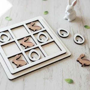 May include: A square wooden tic-tac-toe game board with a white frame and dark grid lines. The game pieces consist of light brown wooden rabbit silhouettes and white oval egg shapes. A small white ceramic rabbit figurine sits nearby.