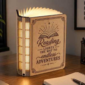May include: A light-up book lamp made of light-colored wood with a spine featuring vertical slats. The cover is engraved with an open book icon and the text: Reading LIGHTS THE WAY TO endless ADVENTURES. The pages emit a warm, soft glow.