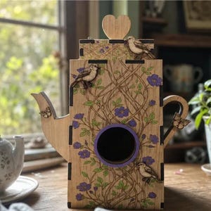 Könnte beinhalten: Ein hölzernes Vogelhaus in Teekannenform mit einer zentralen kreisförmigen Öffnung und einem violetten Rand. Die Außenseite ist mit lasergravierten Ranken, grünen Blättern, violetten Blüten, kleinen Holzvögeln, Schmetterlingen und einem Herz verziert.