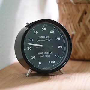 Pode incluir: Um relógio analógico preto com um mostrador verde escuro, algarismos e texto brancos. O relógio tem um design semelhante a um medidor com um ponteiro apontando para a hora atual. O relógio está em uma superfície de madeira com uma cesta tecida ao fundo.