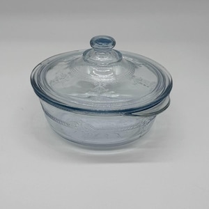 Può includere: Una pirofila in vetro blu chiaro con coperchio abbinato. La pirofila e il coperchio presentano un motivo floreale inciso nel vetro. Il coperchio ha una piccola maniglia rotonda. La pirofila è su una superficie bianca.