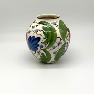 May include: A round ceramic vase with a cream-colored base featuring hand-painted green leaf motifs, blue floral accents, and thin brown swirling lines. The vase has a wide, rounded body and a narrow, circular opening with a dark brown rim.