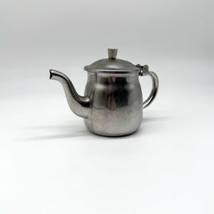May include: A small, brushed metal teapot with a curved spout and a hinged lid. The teapot has a handle and a small knob on the lid. The teapot is made of a silver-colored metal and sits on a white surface.