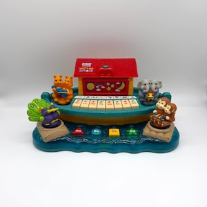 May include: A VTech Land 'n Sea Jamboree musical toy featuring a red roof structure, a piano keyboard with musical notes, and colorful animal figures including a peacock, kangaroos, elephants, and monkeys on a blue base with letter buttons.