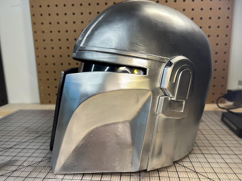 May include: A silver-colored helmet with a smooth, metallic finish. The helmet features a T-shaped visor opening with a dark, recessed interior, a prominent brow ridge, and a raised rectangular ear piece on the side, showcasing a sleek design.