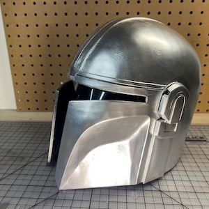 May include: A metallic silver helmet featuring a smooth, rounded dome, a T-shaped visor opening with a dark interior, and a prominent angular faceplate. The helmet has a polished finish with visible structural seams and a mechanical side detail.