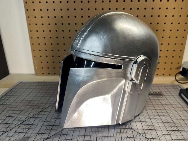 May include: A metallic silver helmet featuring a smooth, rounded dome, a T-shaped visor opening with a dark interior, and a prominent angular faceplate. The helmet has a polished finish with visible structural seams and a mechanical side detail.
