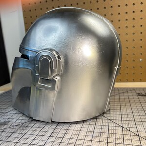 May include: A metallic silver helmet featuring a smooth, rounded dome and a distinct T-shaped visor opening. The side of the helmet displays a raised, rectangular ear piece detail with a vertical slot, finished in a uniform, reflective silver paint.